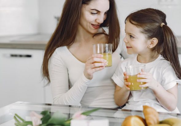 Happy mother and daughter in casual clothes drinking juice and looking at each other while having breakfast together in contemporary kitchen