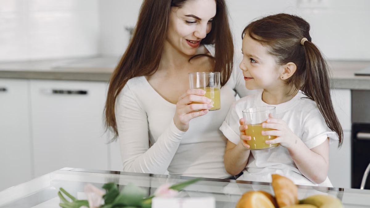 Happy mother and daughter in casual clothes drinking juice and looking at each other while having breakfast together in contemporary kitchen